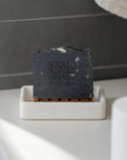 Activated charcoal soap bar on a white soap dish with a dark tiled background, designed for deep cleansing and detoxifying skin.