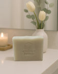 Bar of soap with a logo on a surface, next to a candle and a vase with flowers.