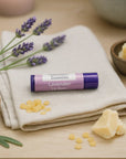 Lavender lip balm tube on beige cloth with beeswax pellets, cocoa butter, lavender sprigs, and natural ingredients on wooden surface