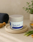 Muscle Relief Body Butter jar on beige plate with turmeric powder, green leaves, and wooden massage tool nearby in spa setting