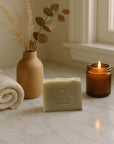 Patchouli & cedarwood moisturizing bar soap with natural ingredients, displayed with rolled towel, dried plants in vase, and lit candle on marble surface