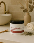 Jar of Patchouli & Cedarwood Body Butter on bathroom counter with towel, wooden spoon, and decorative vase, showcasing natural skincare product.