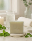 Peppermint bar soap on a white dish with fresh mint leaves, candles, and a glass of water in a bright bathroom setting