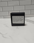 Bar of soap in a black container with a label on a marble surface.