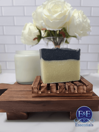 Charcoal and sea clay soap on a wooden soap dish with white roses in the background.