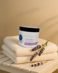 Jar of Lavender Whipped Body Butter on stacked beige towels with fresh lavender sprigs on a wooden surface