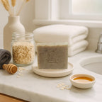 Oatmeal and honey bar soap on a dish with a jar of oats, honey dipper, honey bowl, and folded towels on a bathroom countertop
