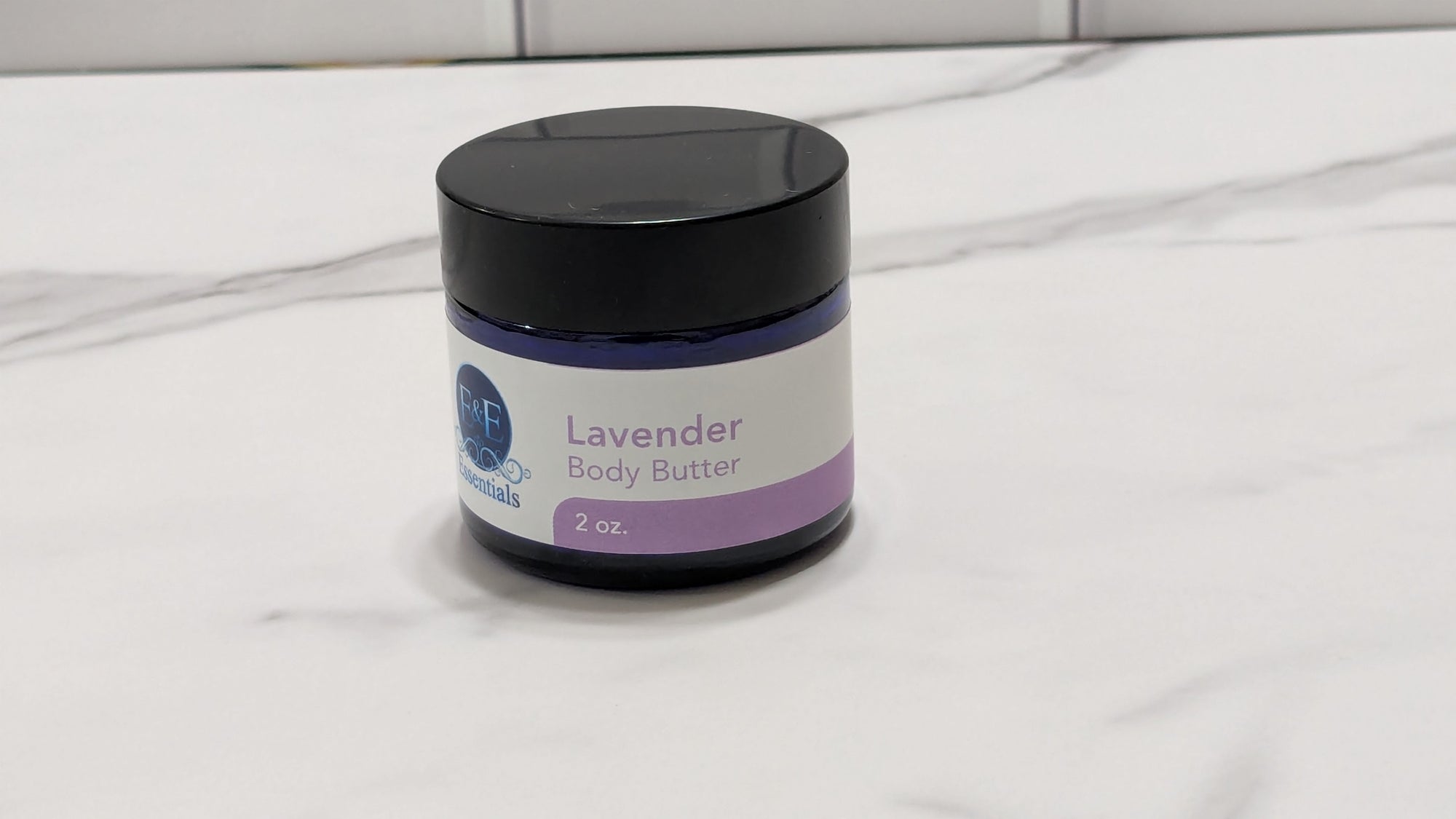 A 2 oz jar of E & E Naturals Lavender Body Butter with a black lid and white/purple label, sitting on a white marble countertop.
