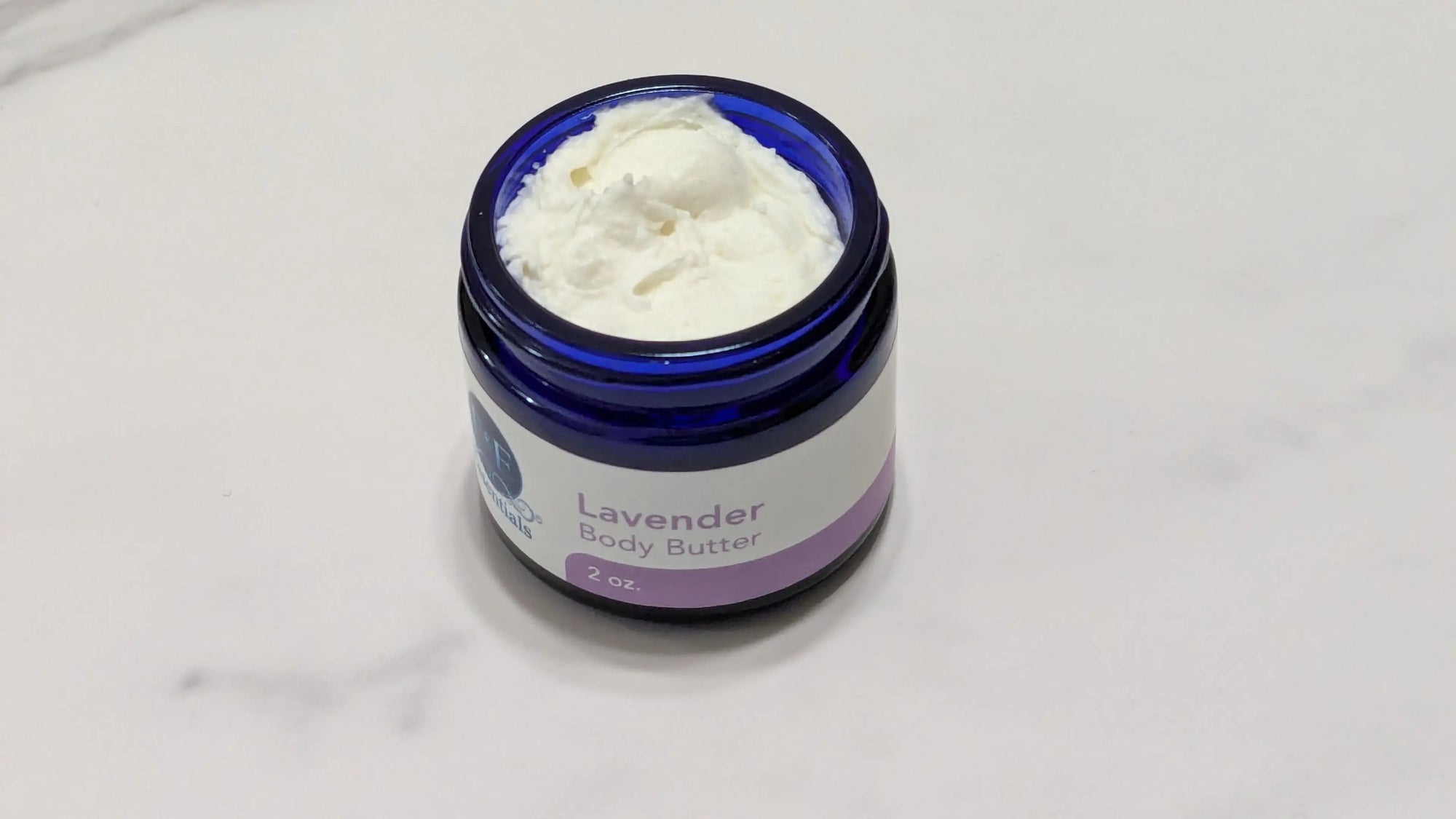 An open jar of lavender body butter with white cream visible inside, set on a light marble-like surface. The jar is dark blue glass with a white and purple label.