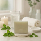Peppermint bar soap on a white dish with fresh mint leaves, candles, and a glass of water in a bright bathroom setting