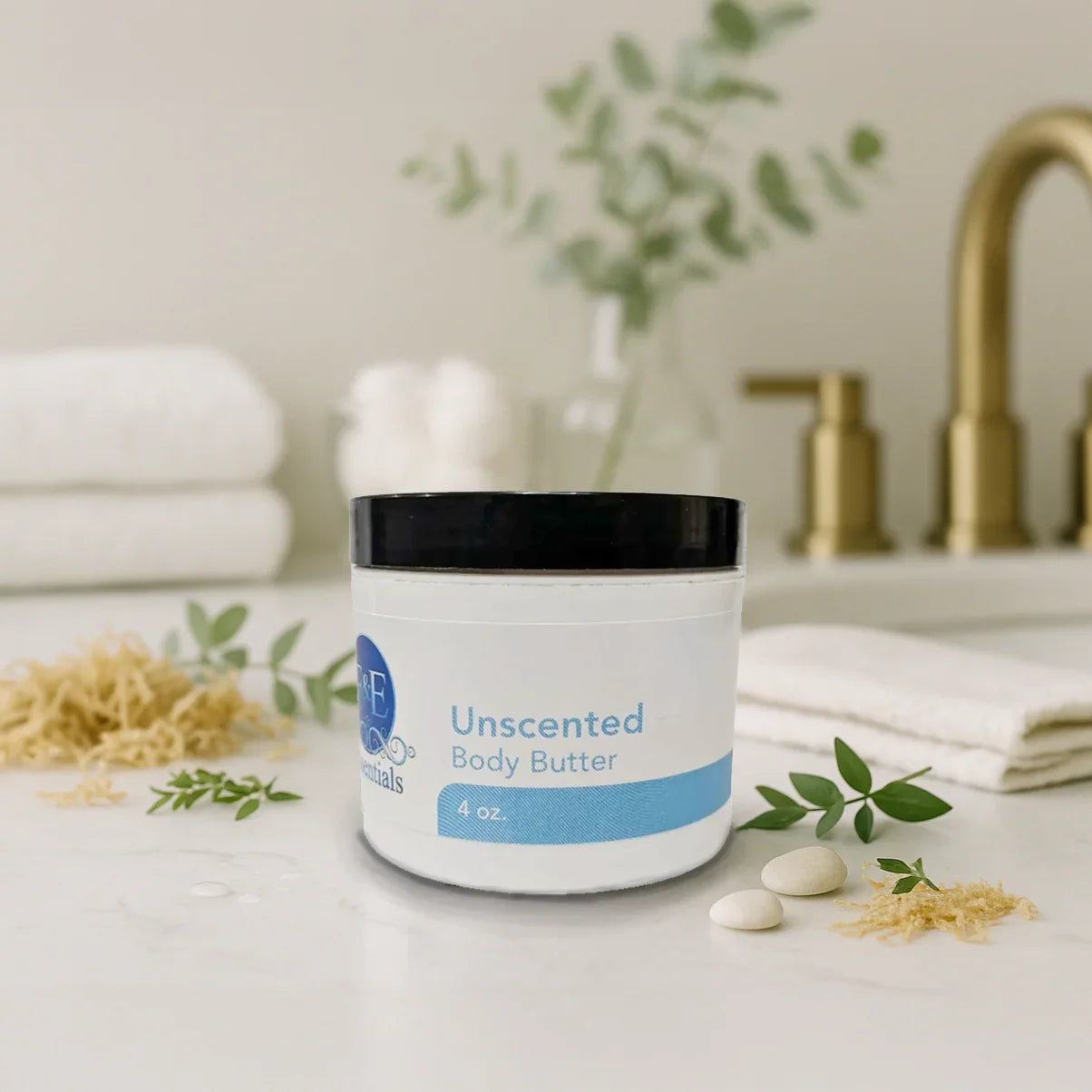 Unscented body butter jar on white countertop with green leaves, white towels, and bathroom accessories in background.