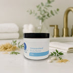 Unscented body butter jar on white countertop with green leaves, white towels, and bathroom accessories in background.