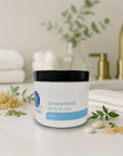 Unscented body butter jar on white countertop with green leaves, white towels, and bathroom accessories in background.